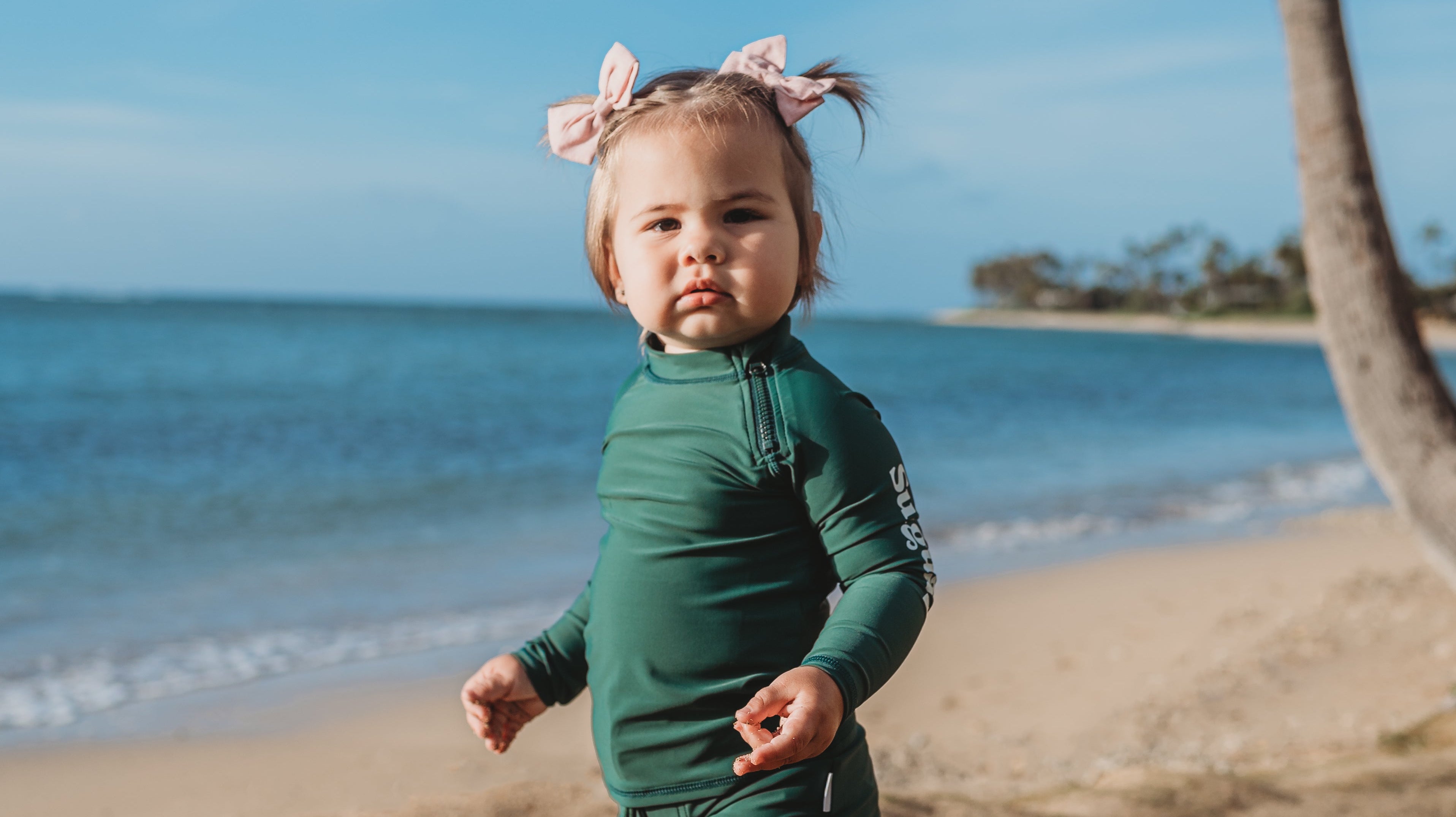 Smiling baby wearing a UPF 50+ sunsuit and sun hat playing on the beach