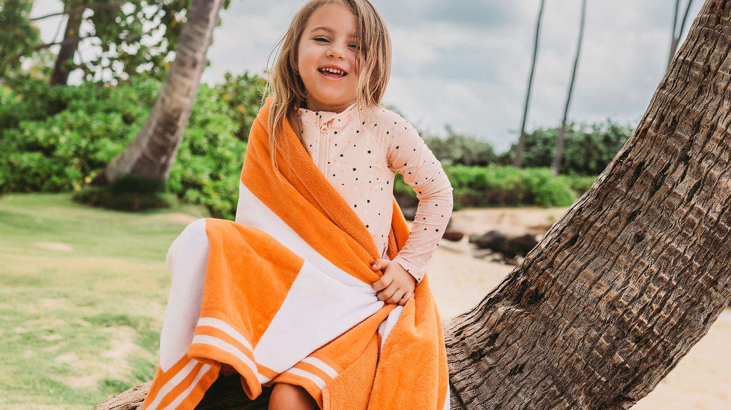 Young girl with an orange towel wearing UPF 50+ swimwear playing on a Hawaiian beach with palm trees in the background.