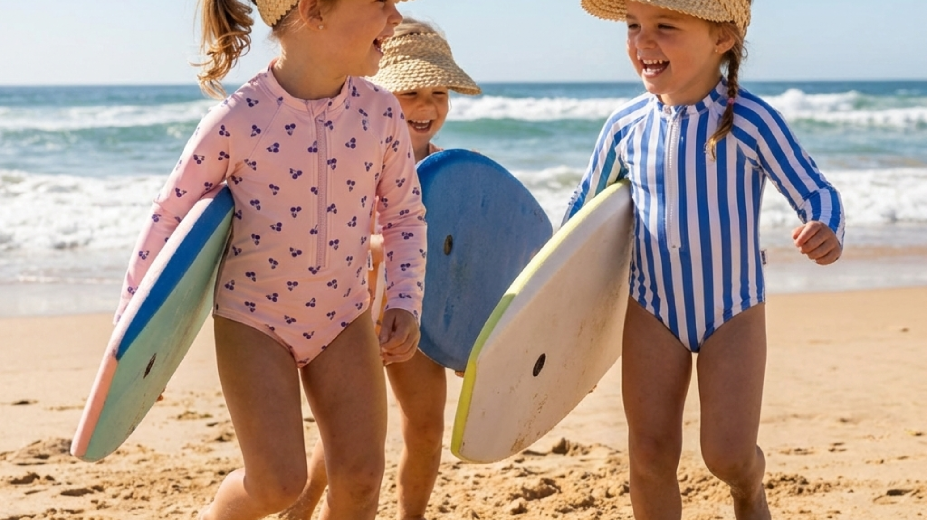 Three young kids in UPF swimsuits and straw sun hats walking on the beach with surfboards