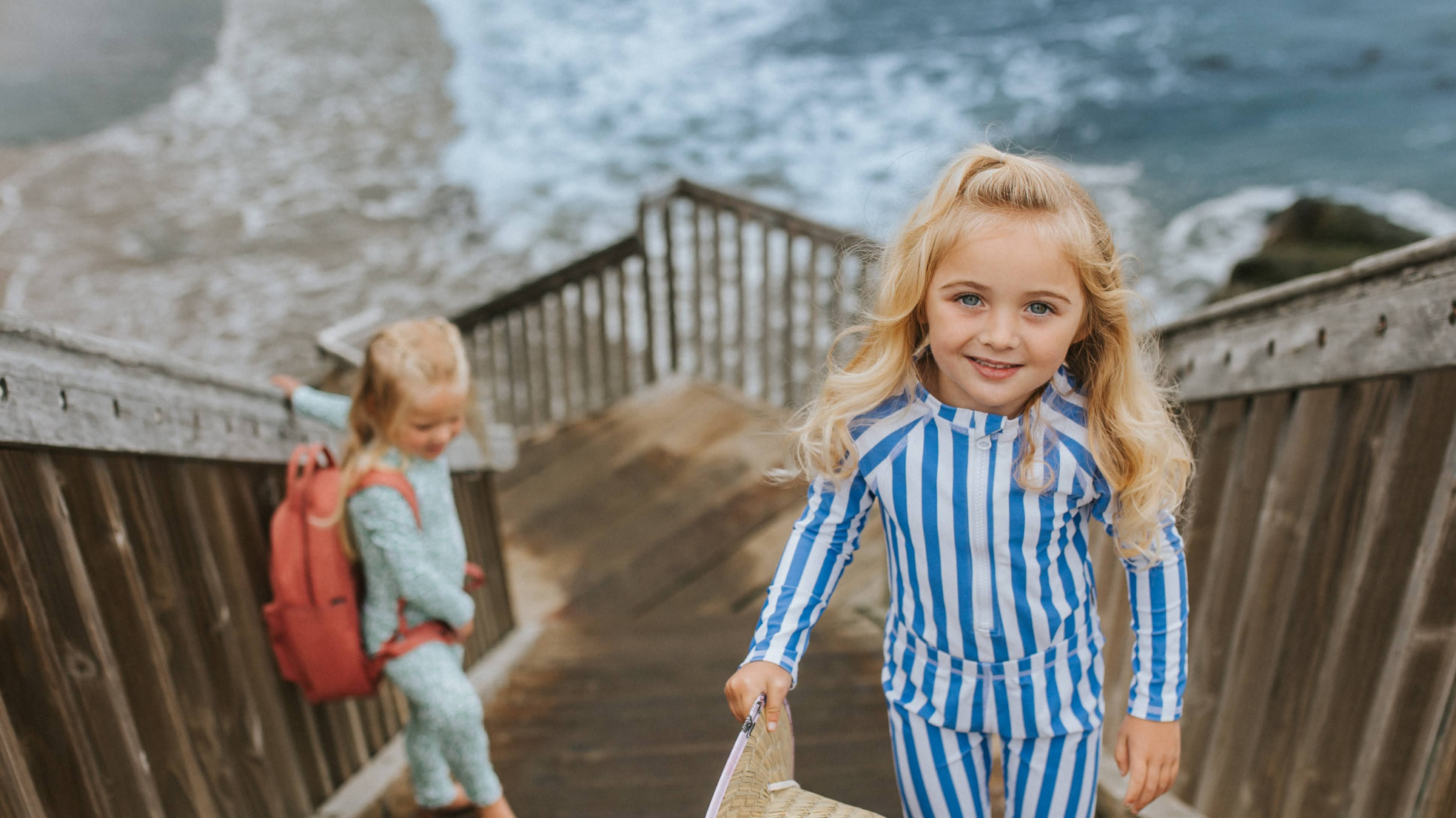 Two young girls wearing long-sleeve UPF swimwear on wooden beach stairs with the ocean in the background.