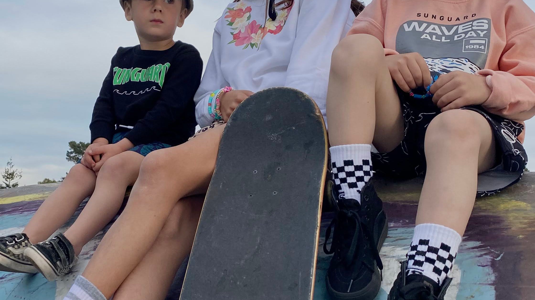 Three kids sitting at a skate park wearing Sunguard sweatshirts and hats, with one holding a skateboard. Casual surf-inspired kids’ style.