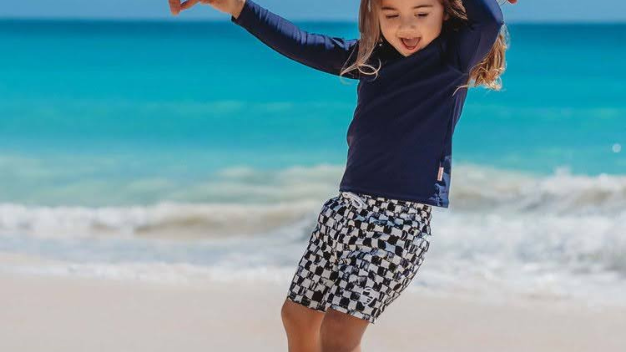 A smiling toddler in a UPF 50+ rashguard playing on the sand on a quiet beach during off-season travel.
