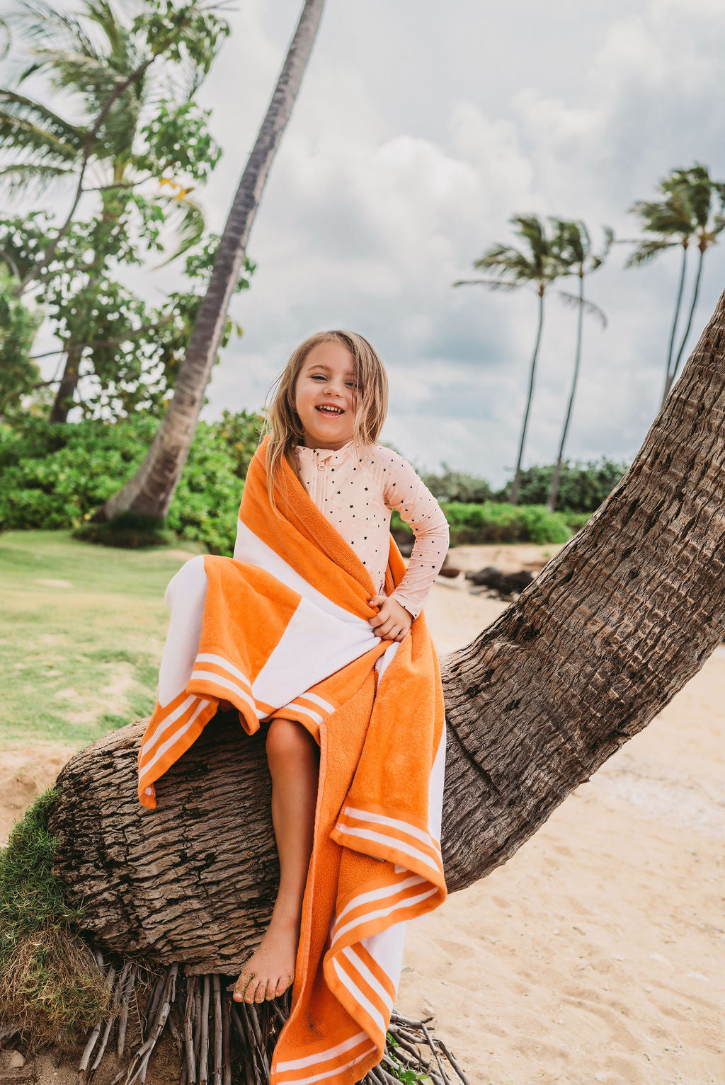 Young girl with an orange towel wearing UPF 50+ swimwear playing on a Hawaiian beach with palm trees in the background.