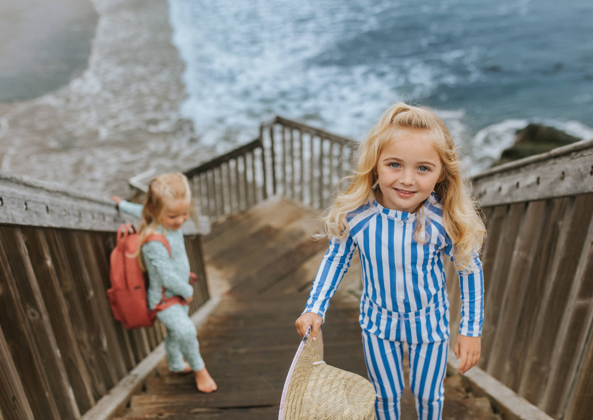 Two young girls wearing long-sleeve UPF swimwear on wooden beach stairs with the ocean in the background.