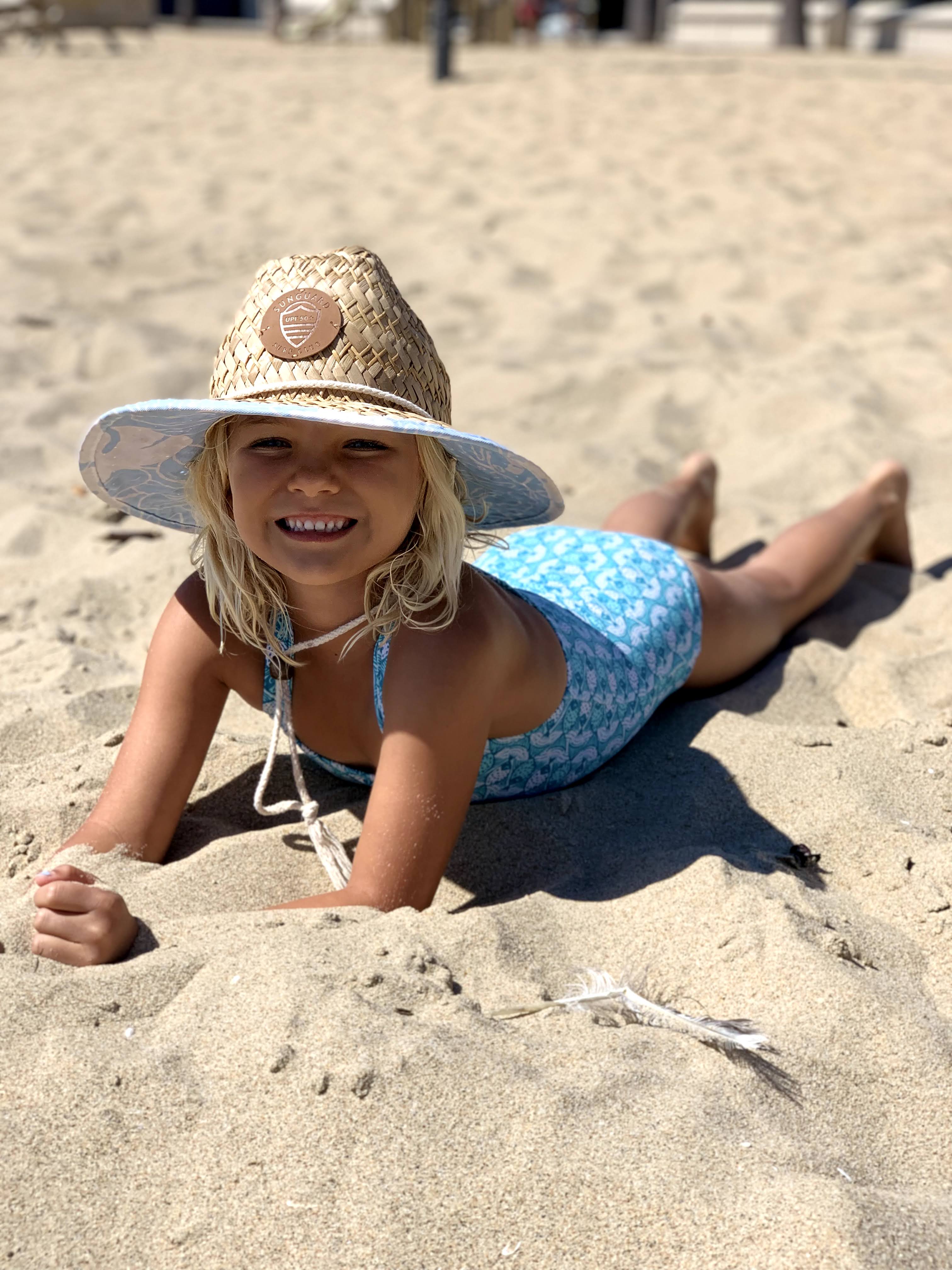 Baby sitting on the sand wearing a straw lifeguard hat with chin strap, dressed in a Sunguard Swim rashguard and shorts at the beach.