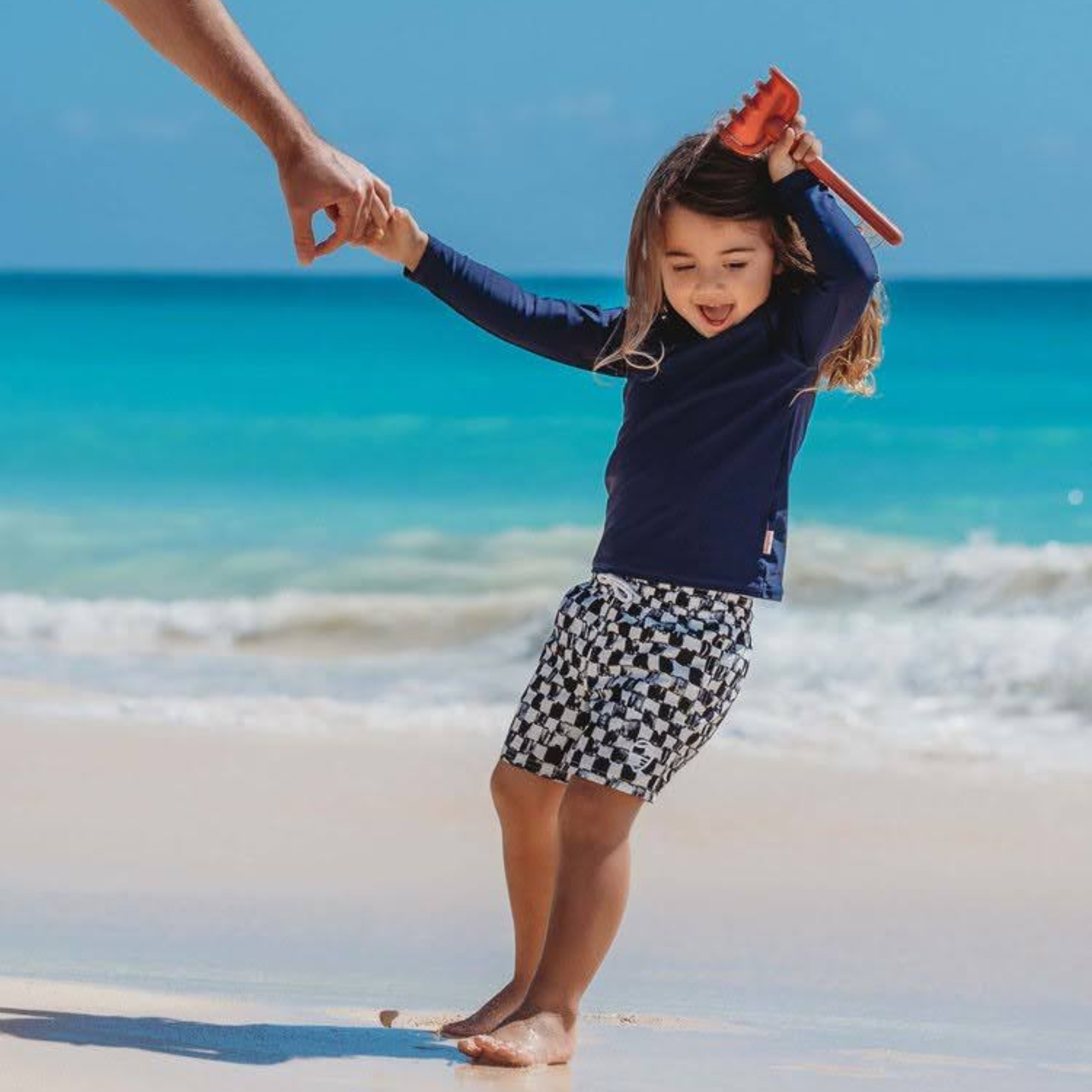 A smiling toddler in a UPF 50+ rashguard playing on the sand on a quiet beach during off-season travel.